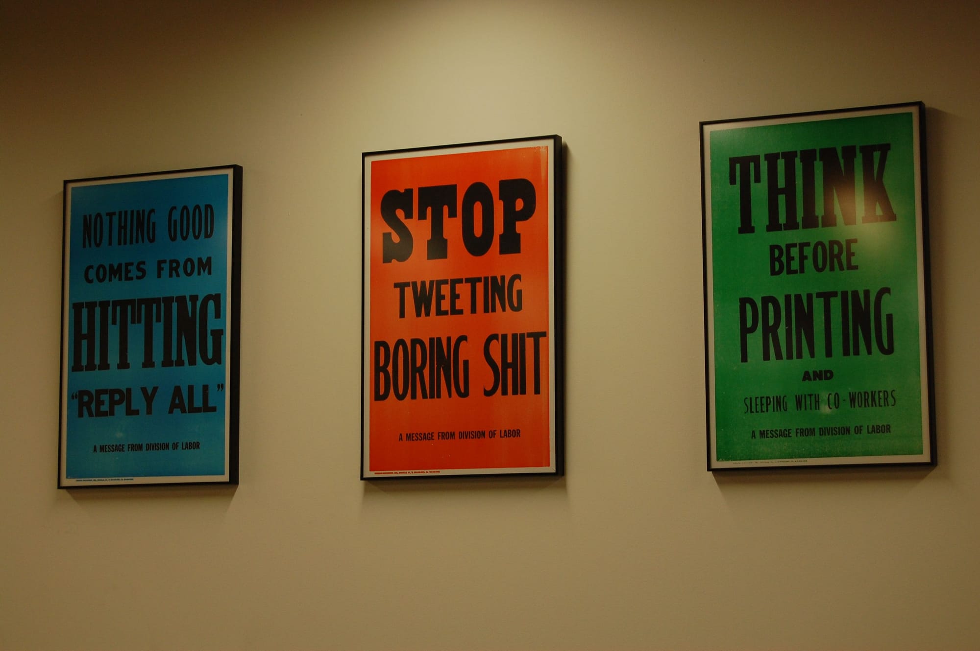 Three colourful posters on a wall displaying humorous workplace messages: “Nothing good comes from hitting reply all,” “Stop tweeting boring shit,” and “Think before printing.”