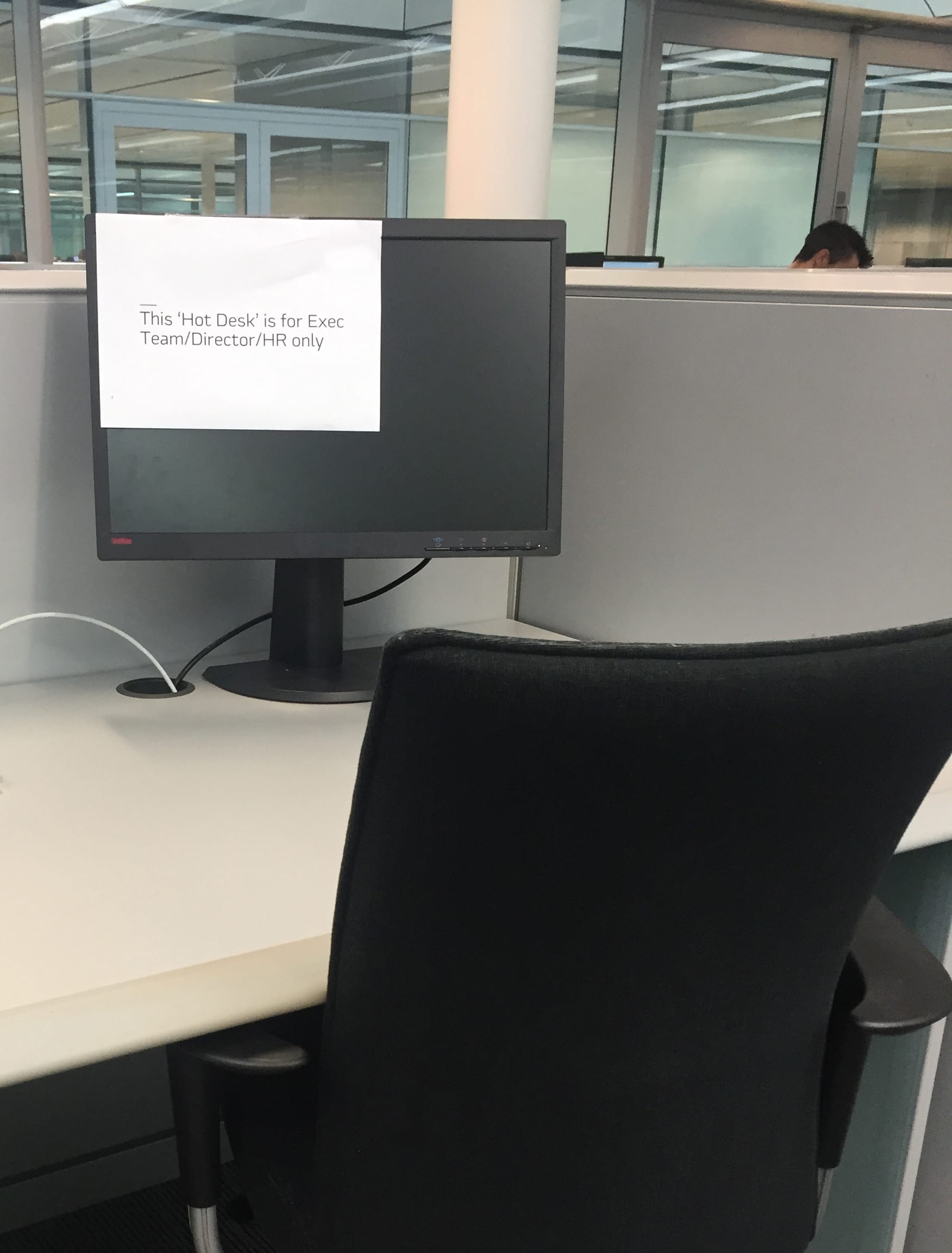 An office workstation with a printed sign taped to the monitor reading “This Hot Desk is for Exec Team / Director / HR only.” A visual cue of hierarchy and exclusion.